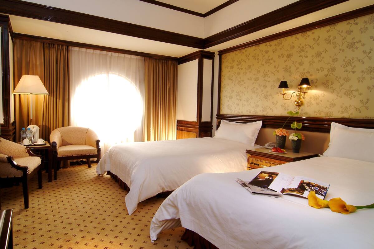A hotel room with two beds, a desk with a lamp, a chair, and a window with curtains.
