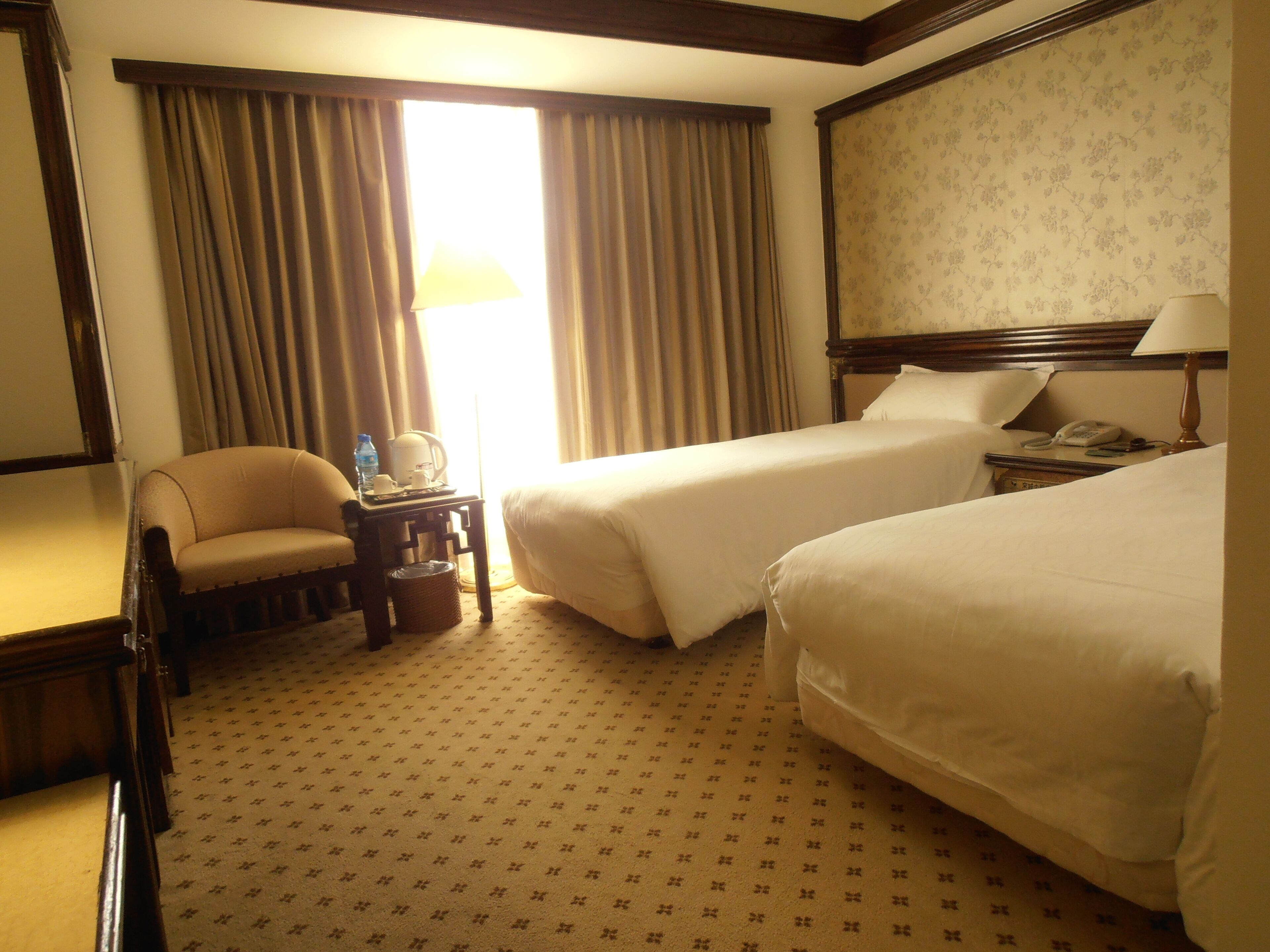 Habitación de hotel con dos camas, una silla, una mesita con lámpara y un ventanal con cortinas.