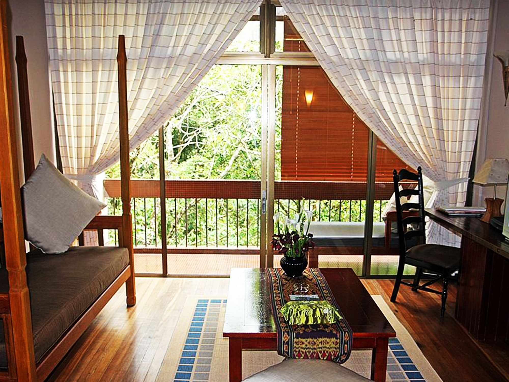 Une pièce avec un canapé en bois, une table recouverte d’une nappe à motifs et une vue sur la verdure à l’extérieur.