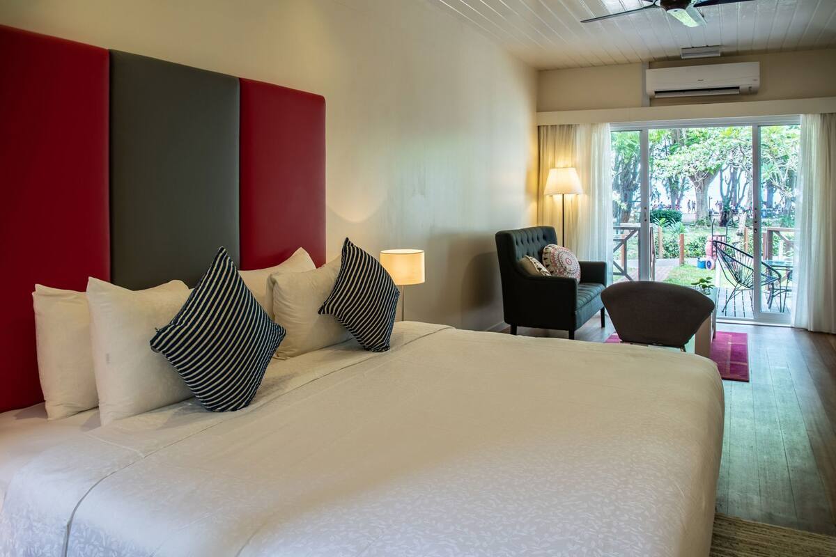 Ein Hotelzimmer mit einem großen Bett, einer Wand mit roten und grauen Akzenten, einem Sessel und Blick ins Freie.