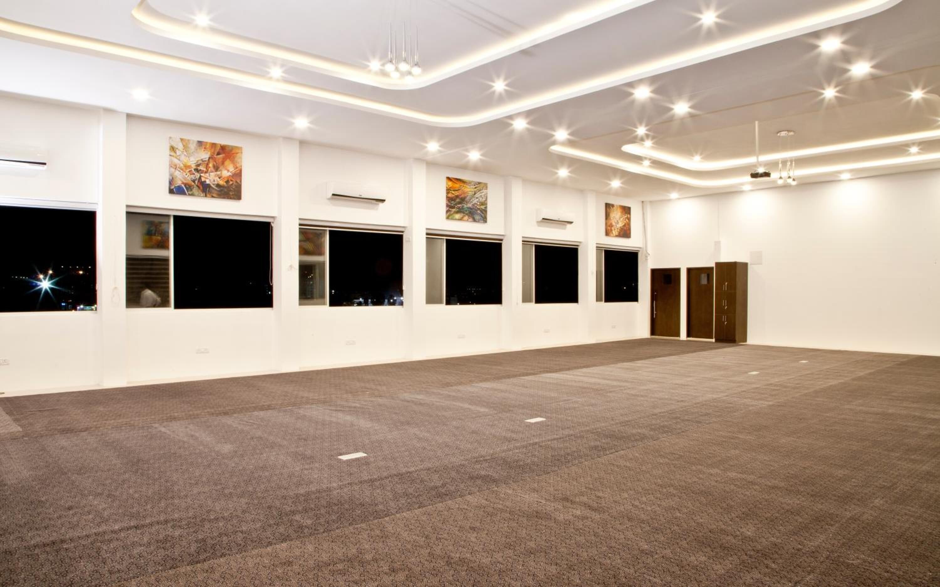 A spacious, well-lit room with large windows, a carpeted floor, and a modern ceiling design.