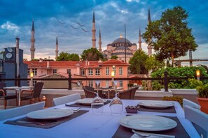 Un espace de restauration aménagé sur le toit, avec des tables pour deux, offrant une vue sur un bâtiment historique doté de plusieurs minarets et d’un dôme.
