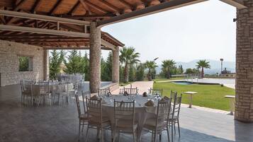 An outdoor dining area with round tables and chairs, stone pillars, and a view of a pool and mountains.