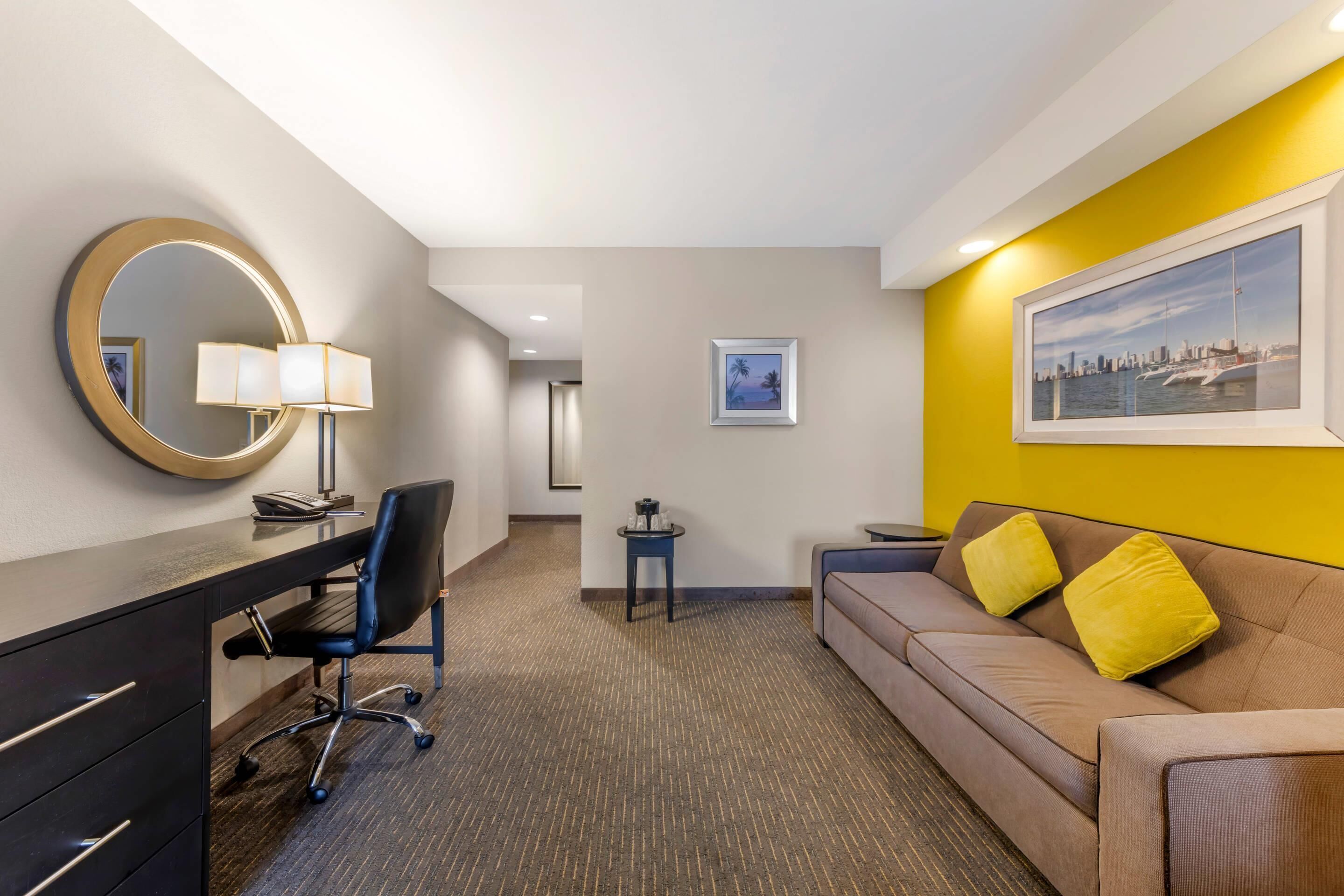 Habitación de hotel con escritorio, silla y sofá con cojines amarillos. Hay un espejo redondo con una lámpara en el escritorio, un cuadro en la pared y una mesita de noche con lámpara.