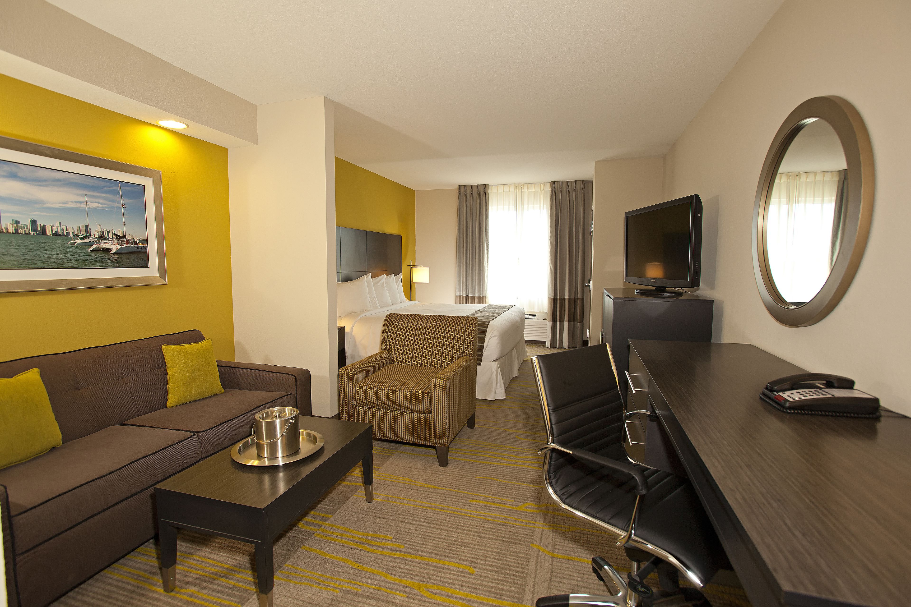 A hotel room with a sofa, armchair, dining table, TV, and a large mirror.