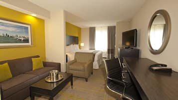 A hotel room with a sofa, armchair, dining table, TV, and a large mirror.