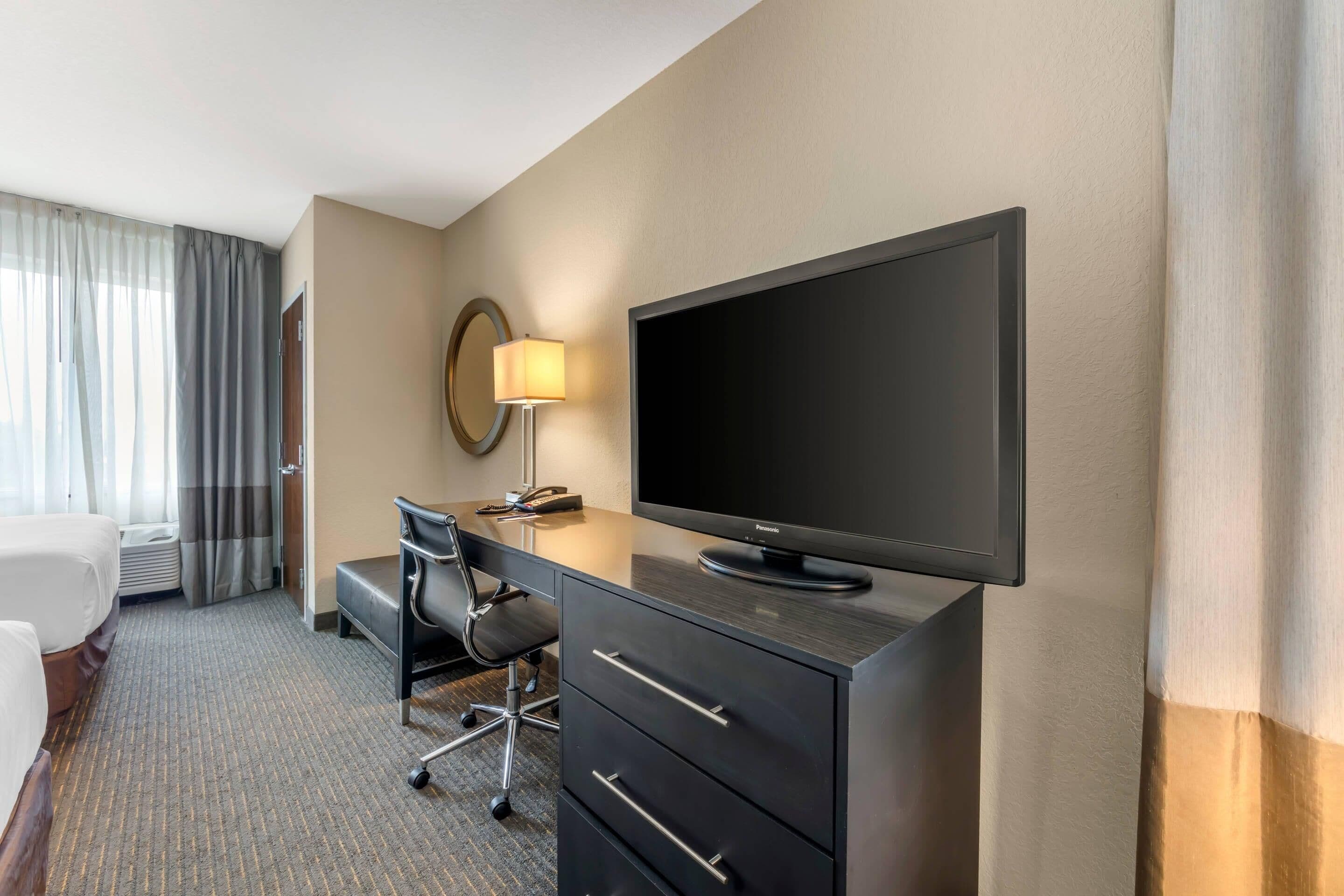 Une chambre d’hôtel comprenant un lit, un bureau avec un téléviseur à écran plat, une chaise, une lampe et une fenêtre avec des rideaux.
