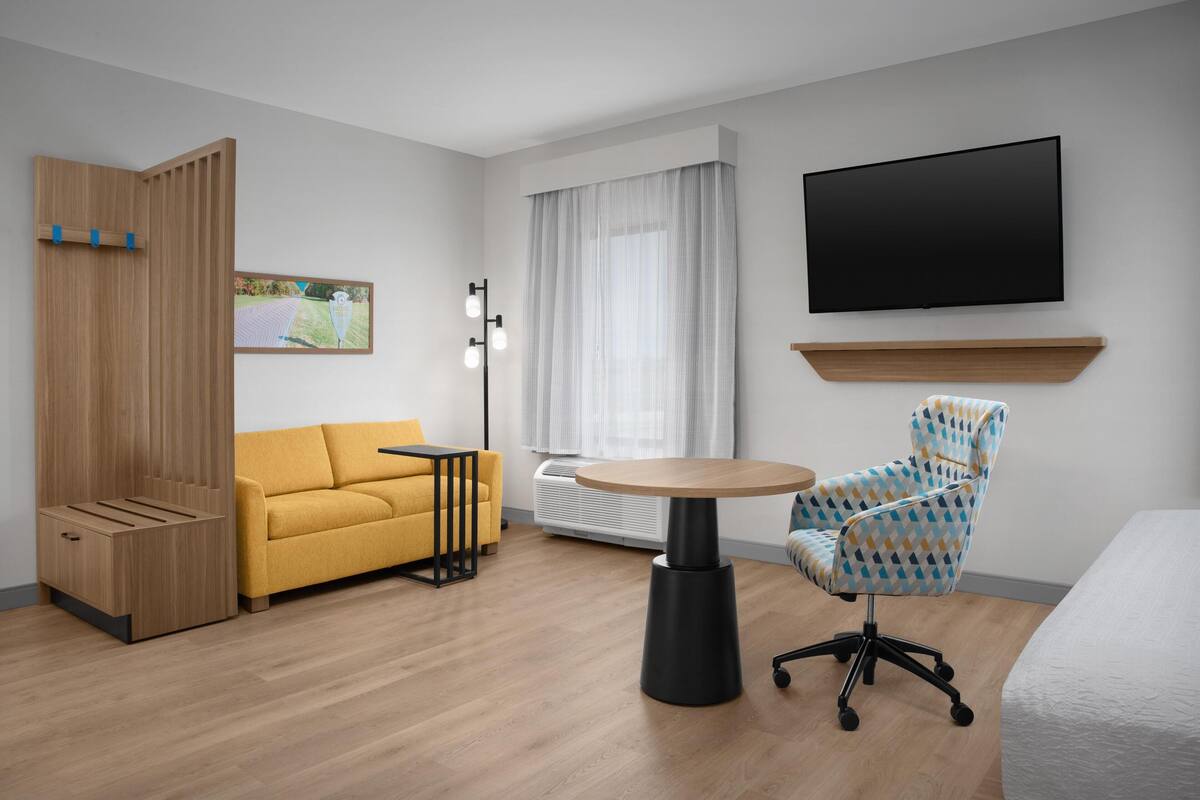 A modern hotel room with a wooden wardrobe, a yellow sofa, a round table, a patterned armchair, a flat-screen TV, and a large window with curtains.