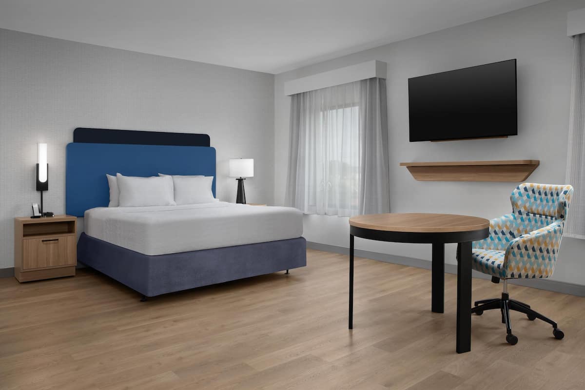 A modern hotel room with a bed, bedside lamps, a TV, a small table, and a patterned armchair.
