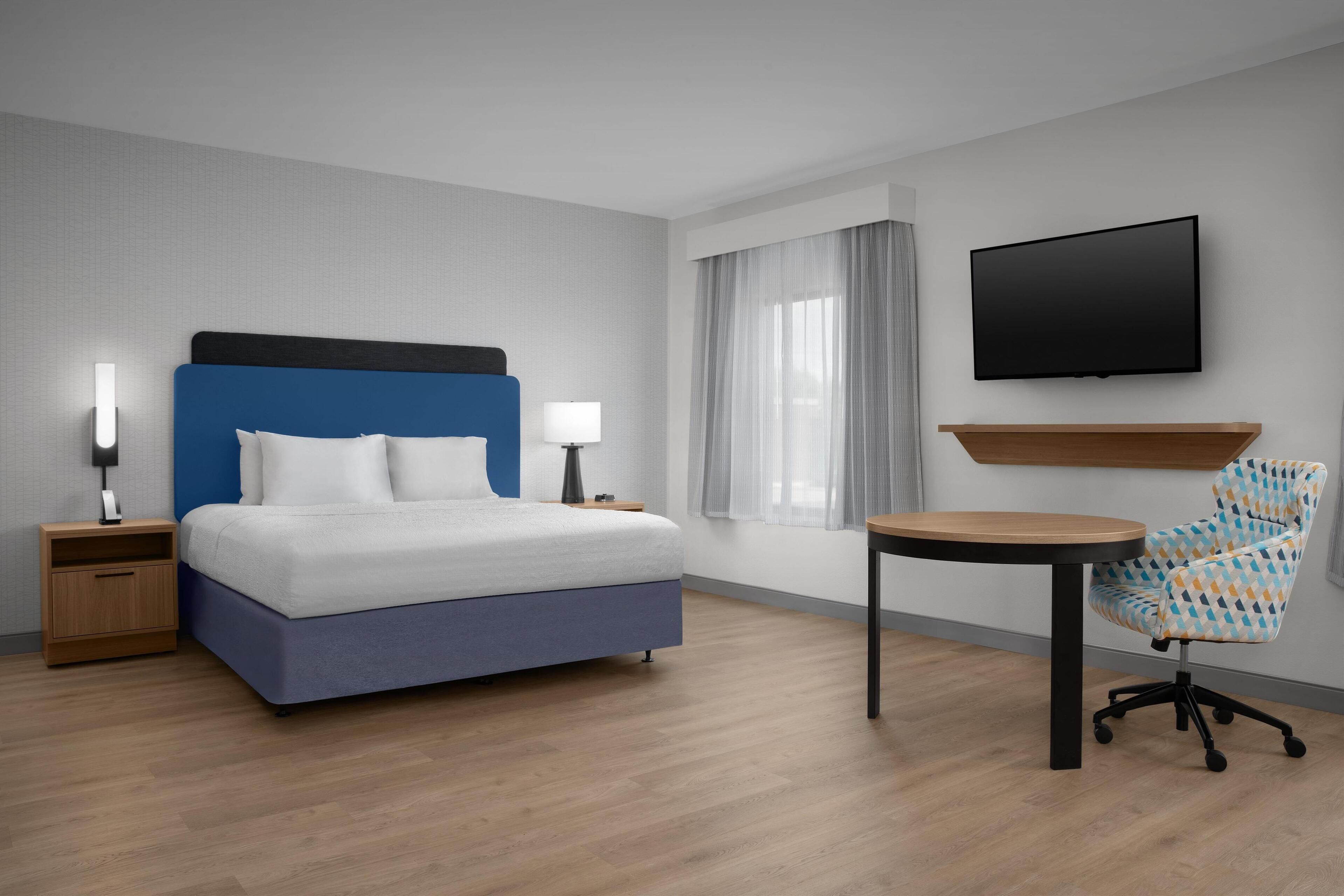 Une chambre d’hôtel avec un lit, des tables de chevet, une télévision, une chaise et une fenêtre avec des rideaux.