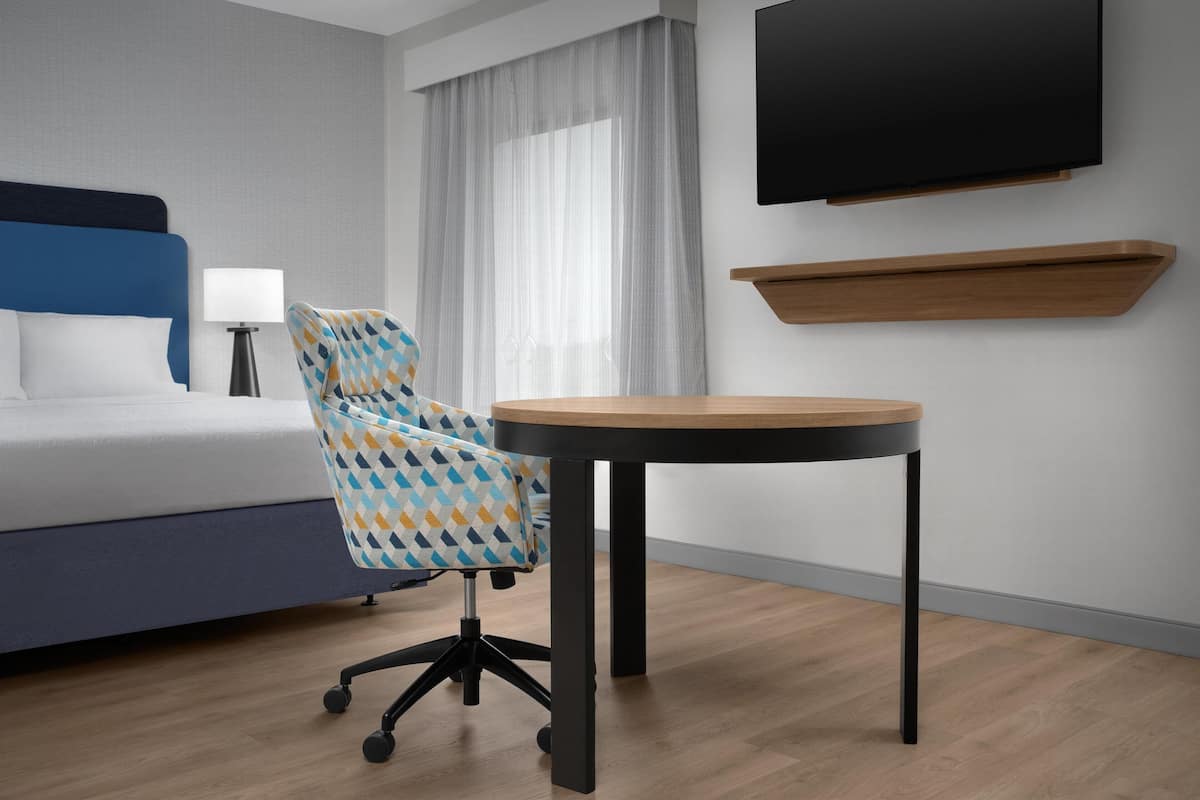 A modern hotel room with a bed, a desk, a chair, a TV, and a window with curtains.