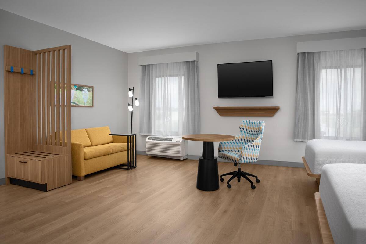 A hotel room with a wooden wardrobe, a yellow sofa, a round table, a patterned armchair, a TV, and two beds.