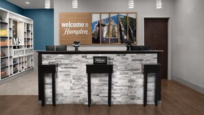 Een receptiebalie met een bordje 'Welcome to Hampton', een 'Hilton Honors' plaquette en een uitzicht op een gebouw door een raam.