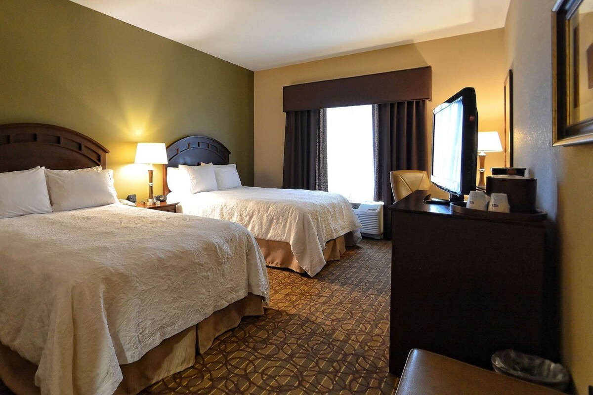 A hotel room with two beds, a desk, a TV, and a window with curtains.