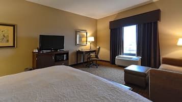 A hotel room with a large bed, a desk with a chair, a television, an air conditioning unit, and a window with curtains.