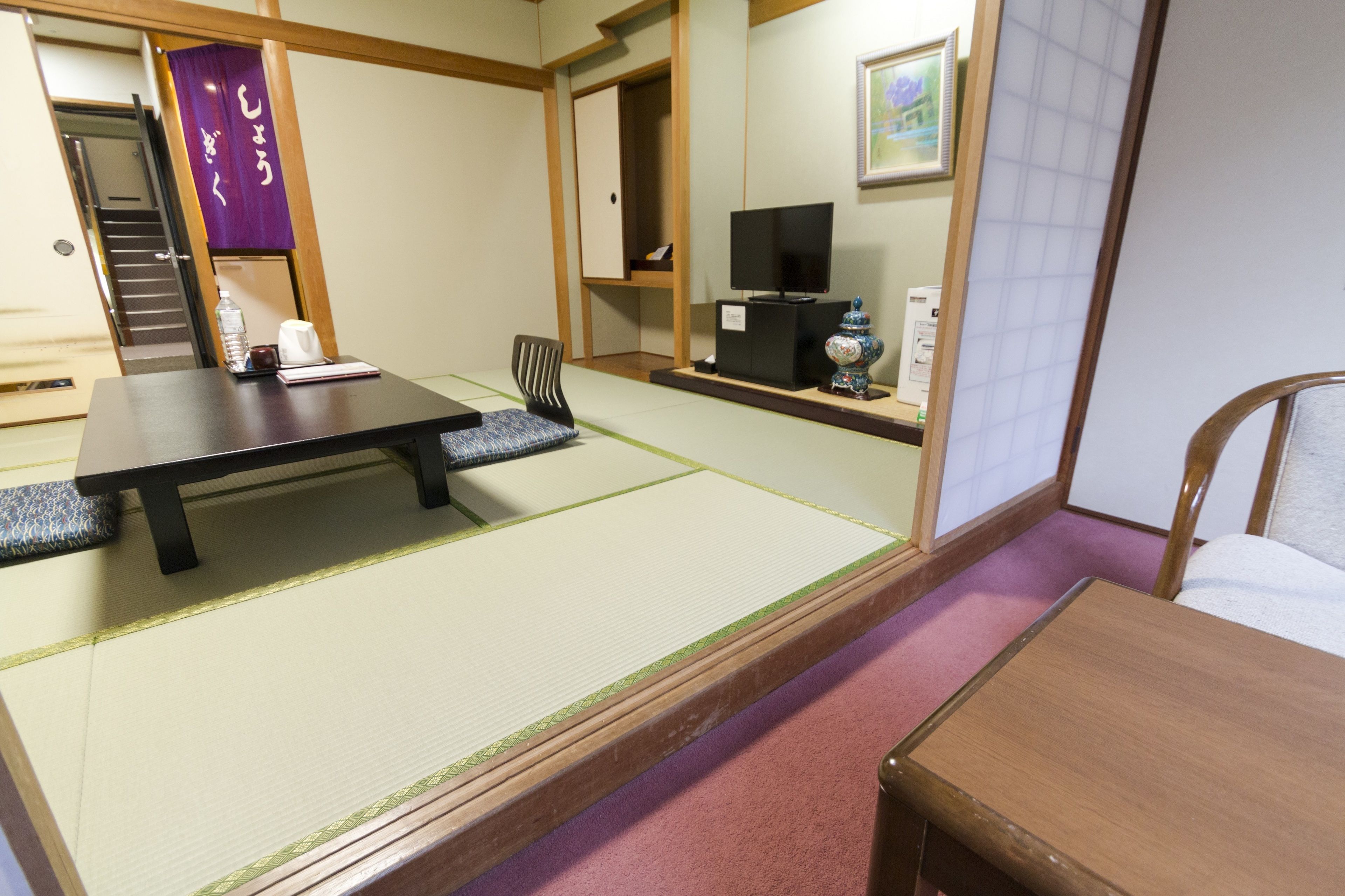 Una habitación con esteras de tatami, una mesa baja, una silla, un televisor sobre un soporte y una estantería con objetos.