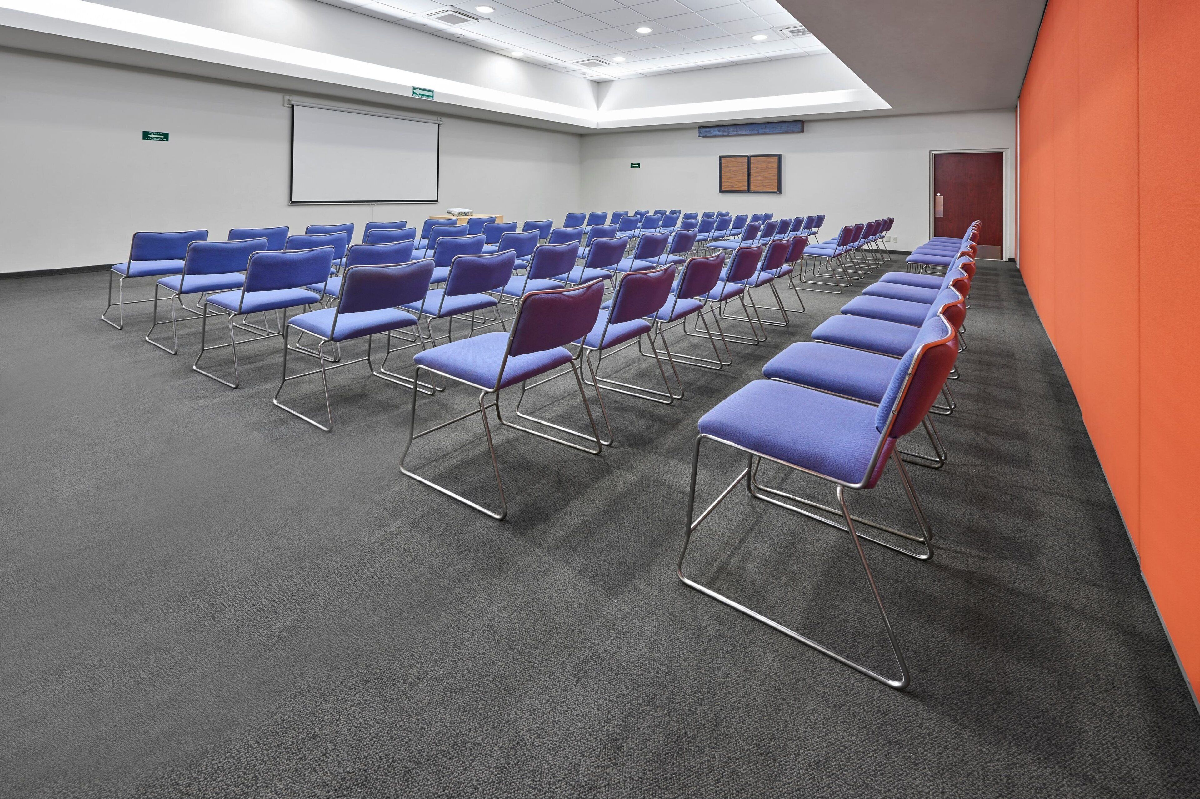 A conference room with rows of blue and red chairs, a whiteboard, and a projector screen.