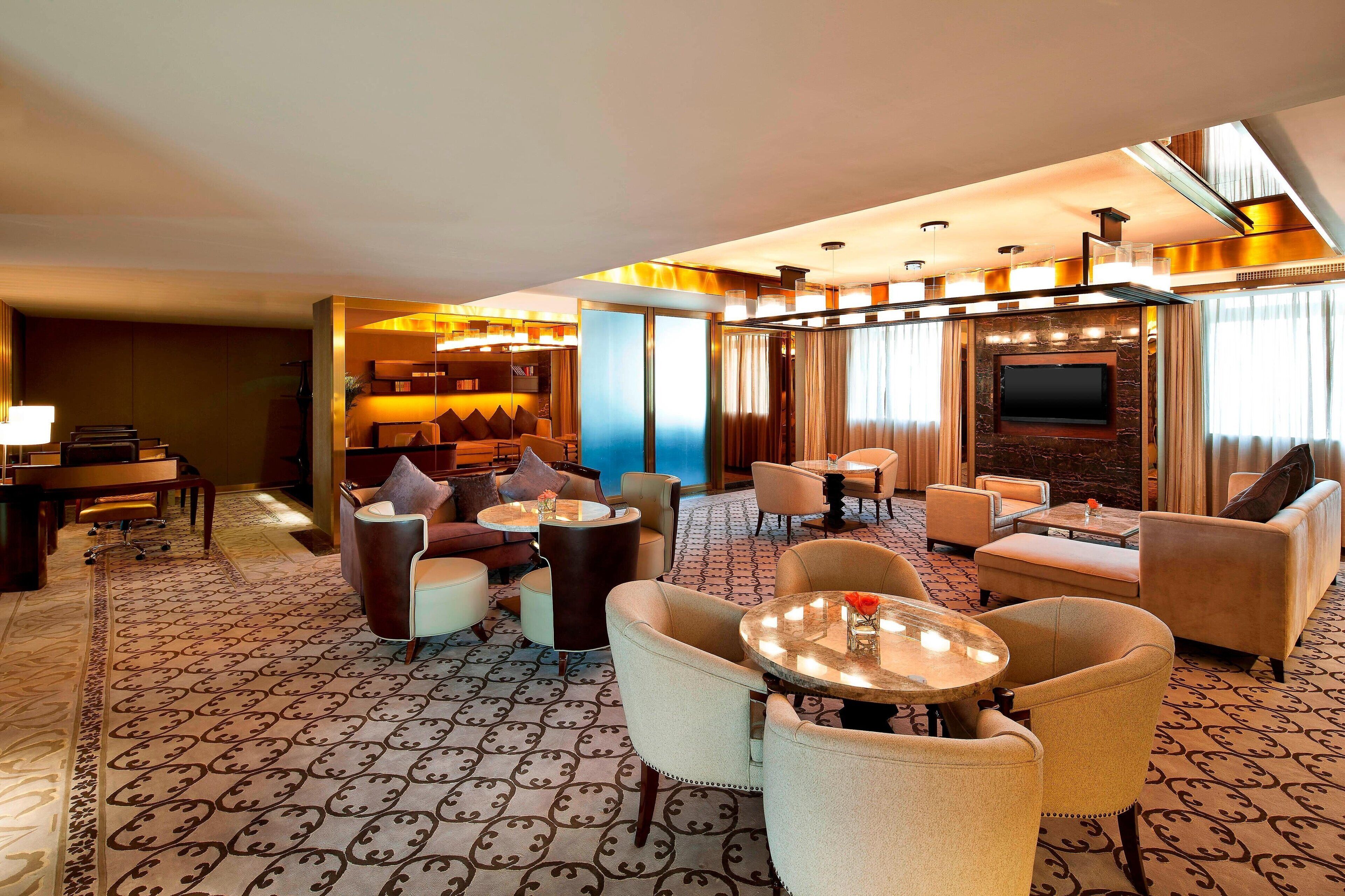 A hotel lobby with a patterned carpet, multiple seating areas including sofas and chairs, a television mounted on a wall, and a bar area with a counter and stools.