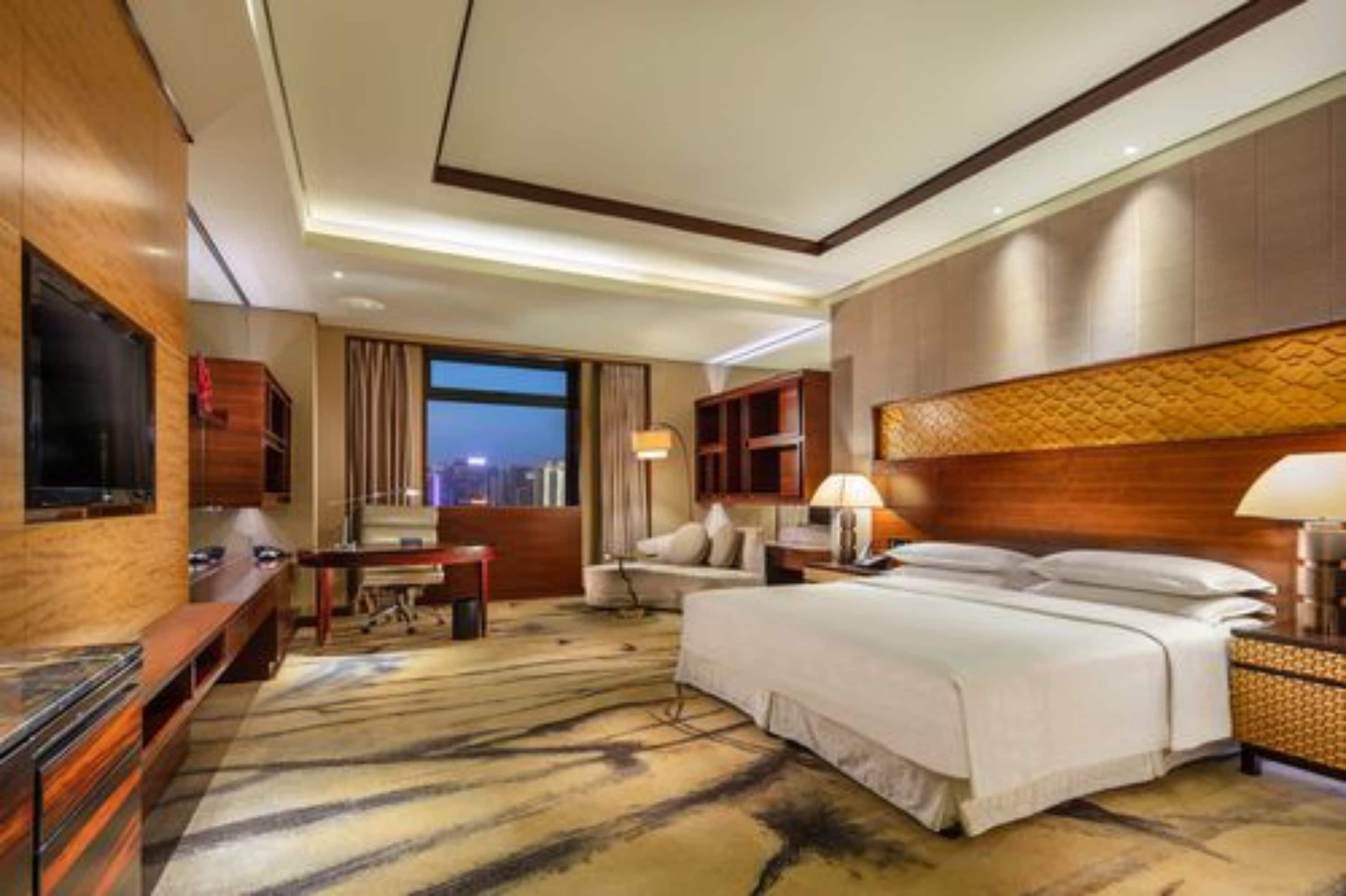 A hotel room with a large bed, a TV, a desk, and a view of the city at dusk.