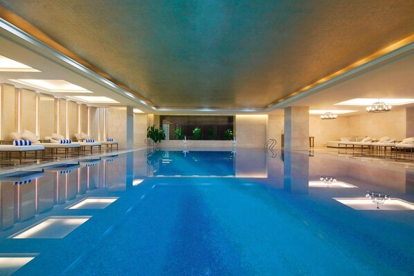 A large indoor swimming pool with lounge areas on both sides, a clear ceiling, and chandeliers.