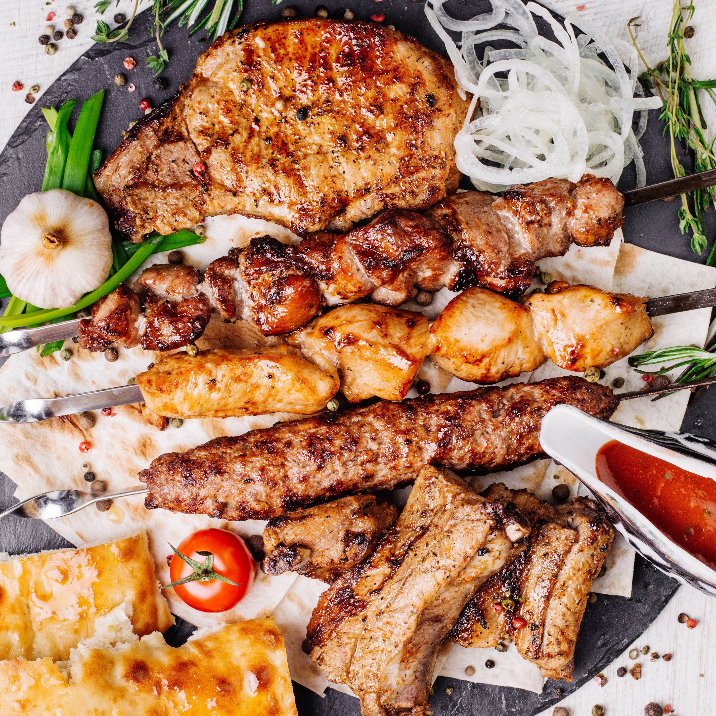 Variedad de carnes a la parrilla, incluyendo brochetas y costillas, servidas en una tabla de pizarra con guarniciones como cebollas y hierbas aromáticas.