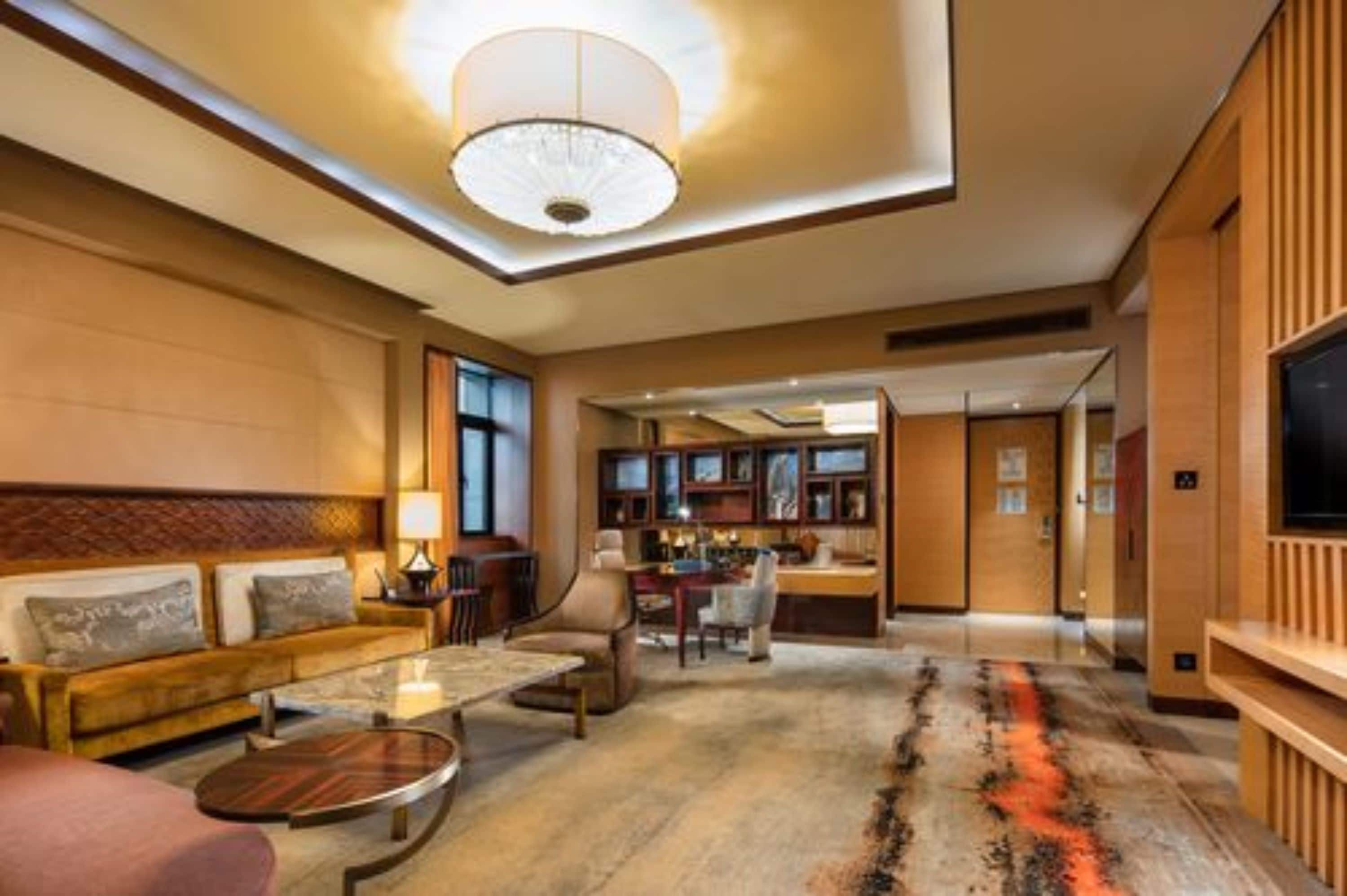 A spacious living area with a sofa, coffee table, and a television. There is a ceiling light fixture and a patterned rug on the floor.