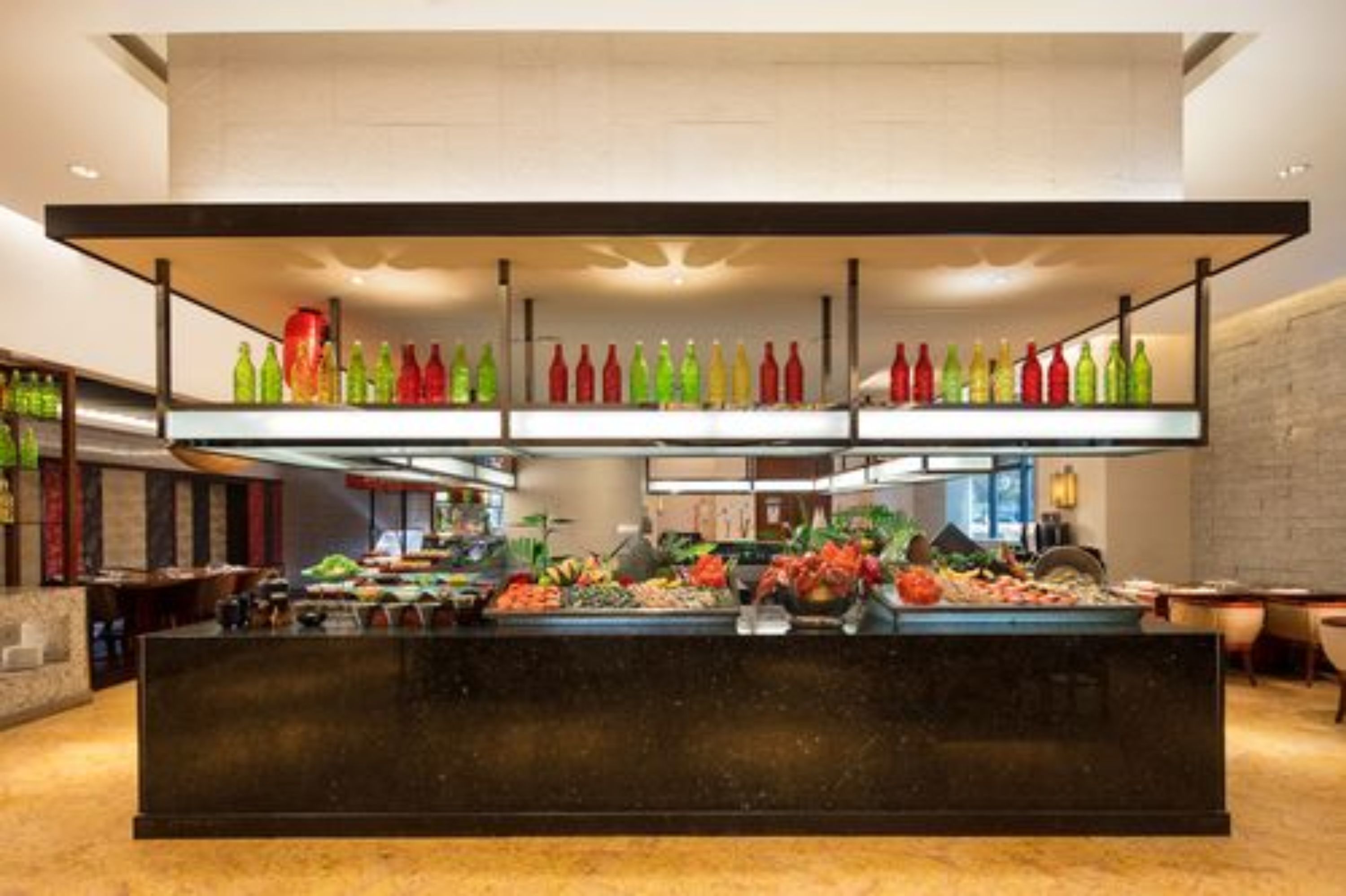 A hotel lobby with a central reception desk, a fruit and vegetable display, and shelves with colorful bottles.