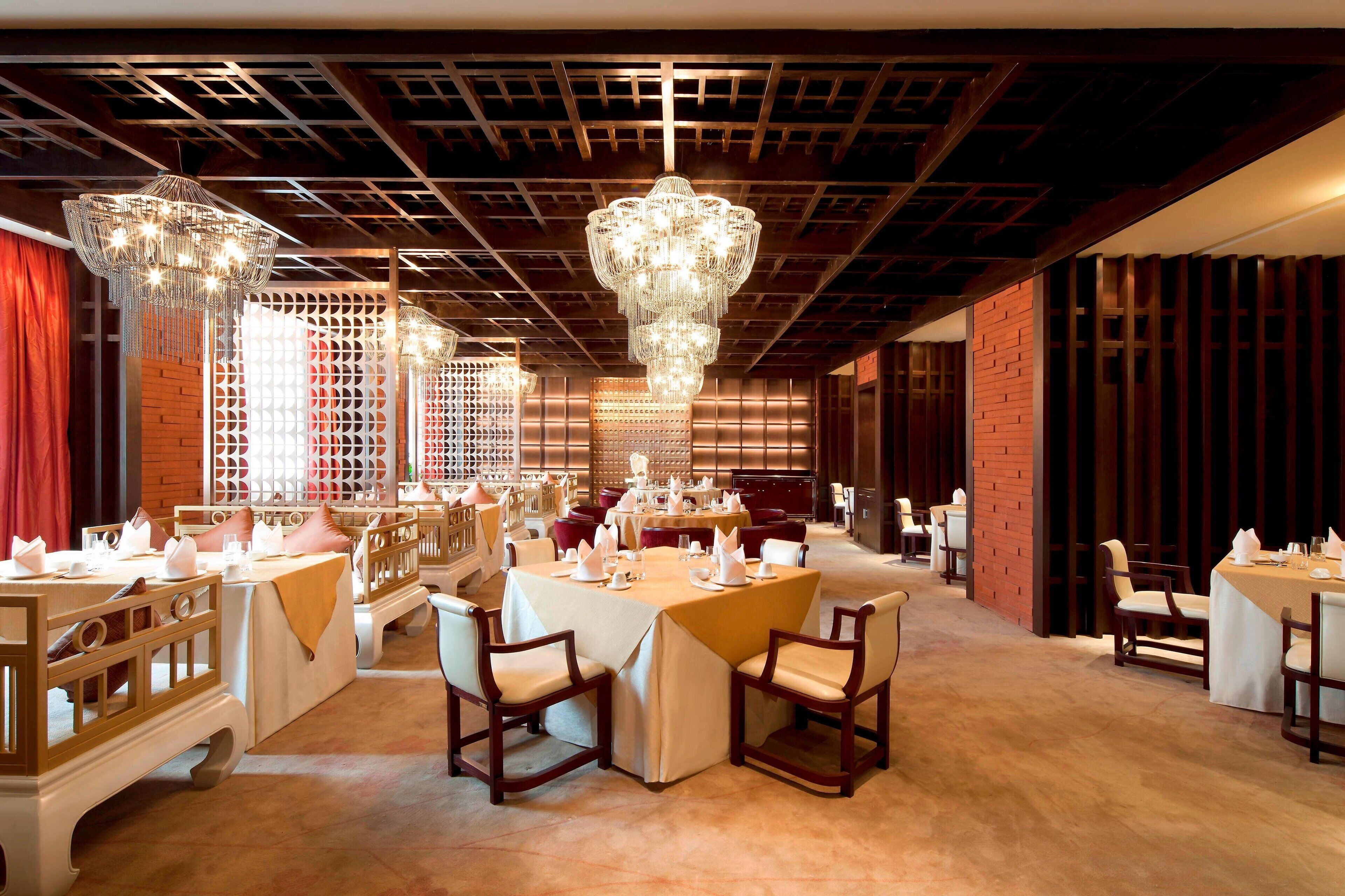 Een ruime eetruimte met meerdere tafels gedekt voor een maaltijd, elegante kroonluchters en een houten plafondconstructie.
