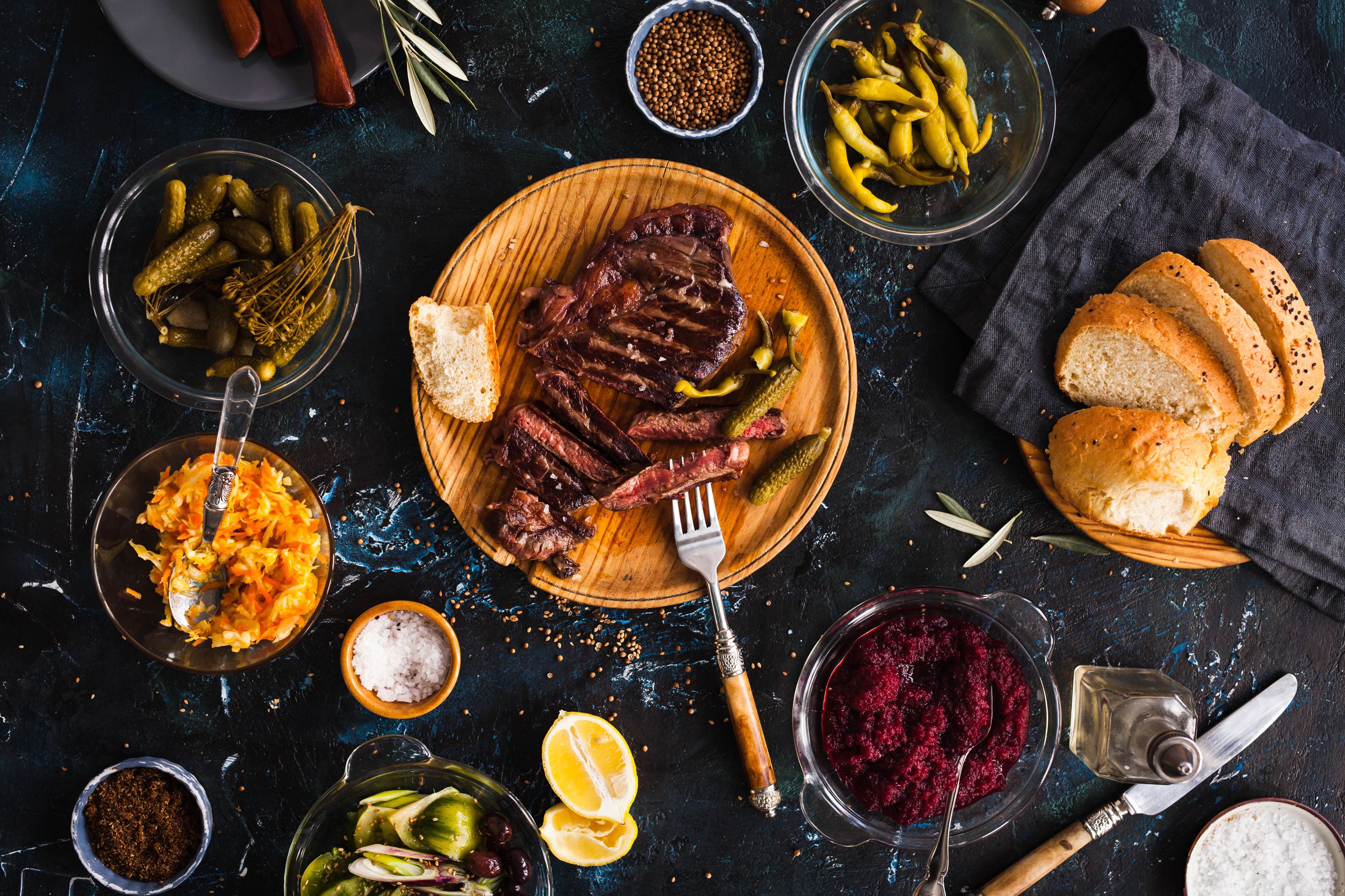 A wooden board with sliced meat, pickles, and bread on a dark surface.