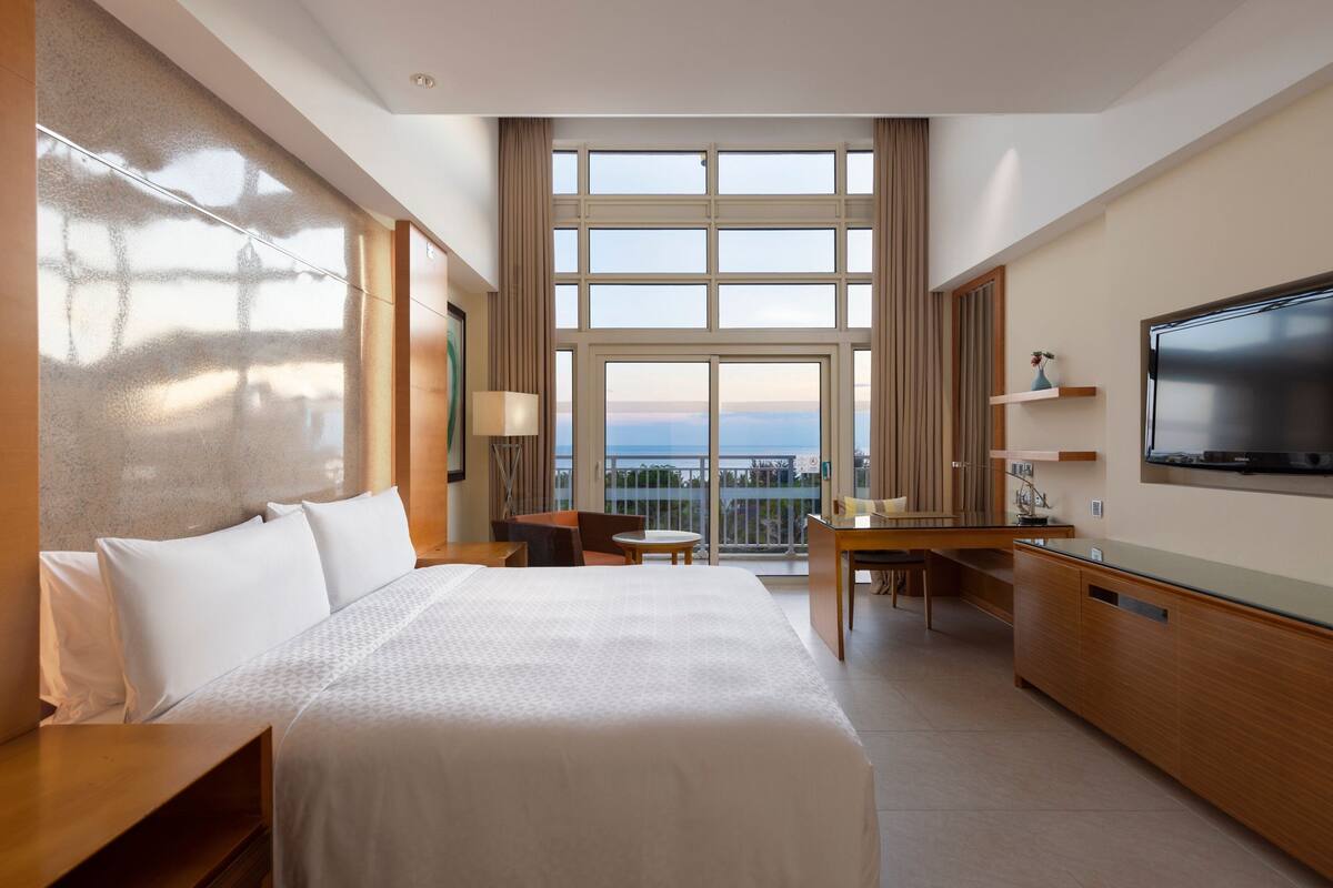 Habitación de hotel con una cama grande, un escritorio, una silla, un televisor y un balcón con vistas.