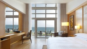 Habitación de hotel con una cama grande, un escritorio, una silla, un balcón con vistas al mar y dos lámparas de noche.