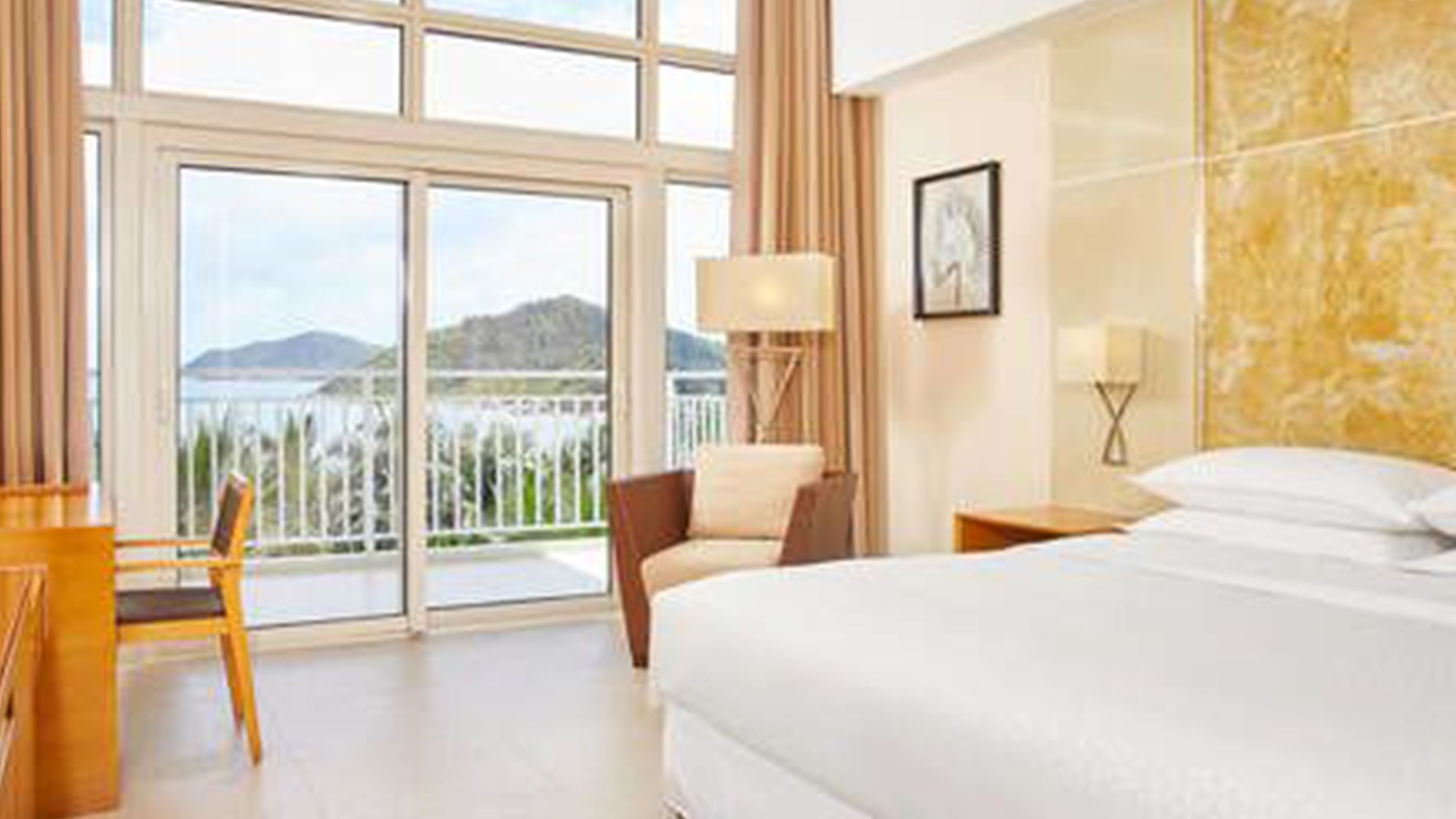 A hotel room with a large bed, a chair, a nightstand, a lamp, and a view of a body of water and mountains through large windows.