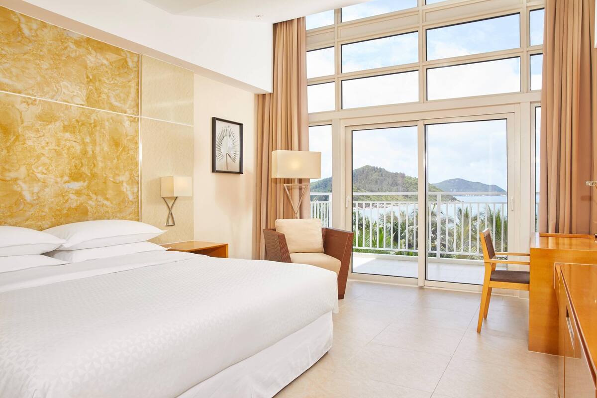 Habitación de hotel con una cama grande, un escritorio, una silla, una lámpara y vistas al mar a través de amplios ventanales.