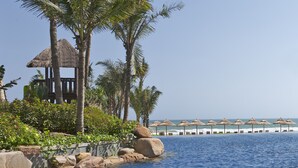 Un área junto a la piscina con una estructura de techo de paja, palmeras y sombrillas de playa.