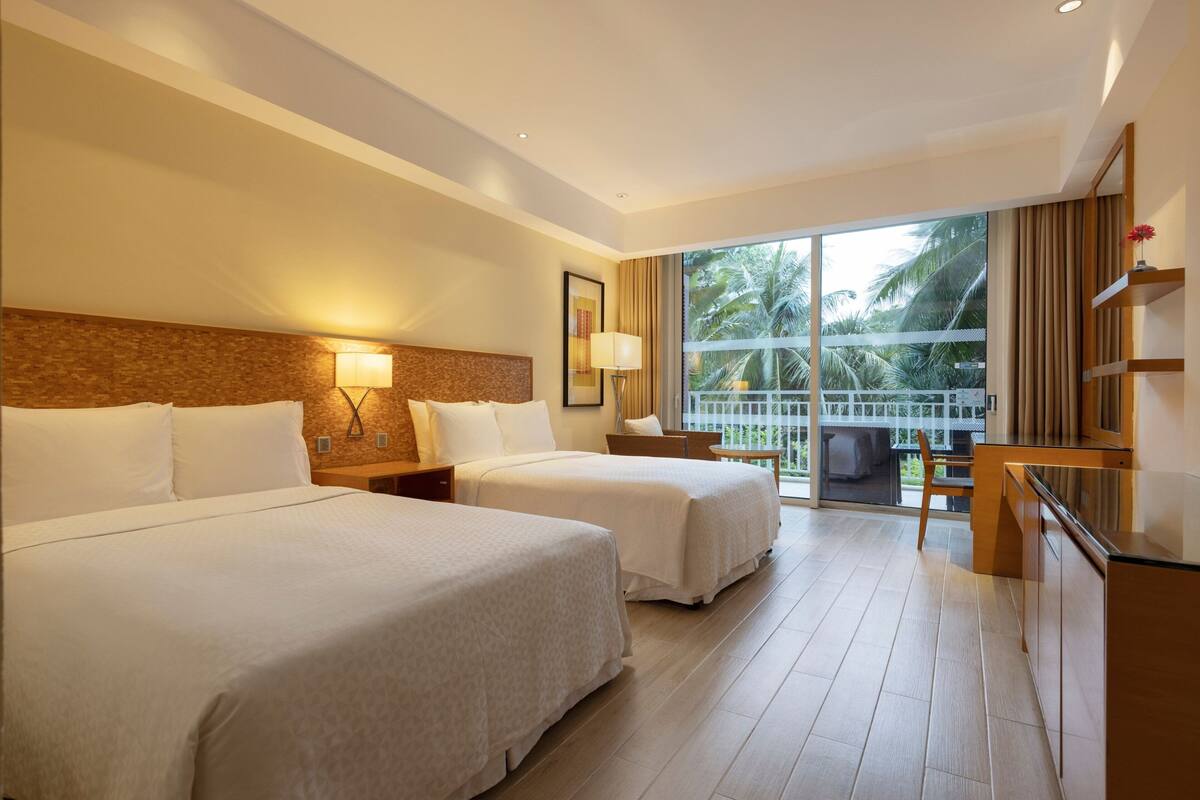 Habitación de hotel con dos camas, un balcón y vistas a palmeras, un escritorio y una silla.