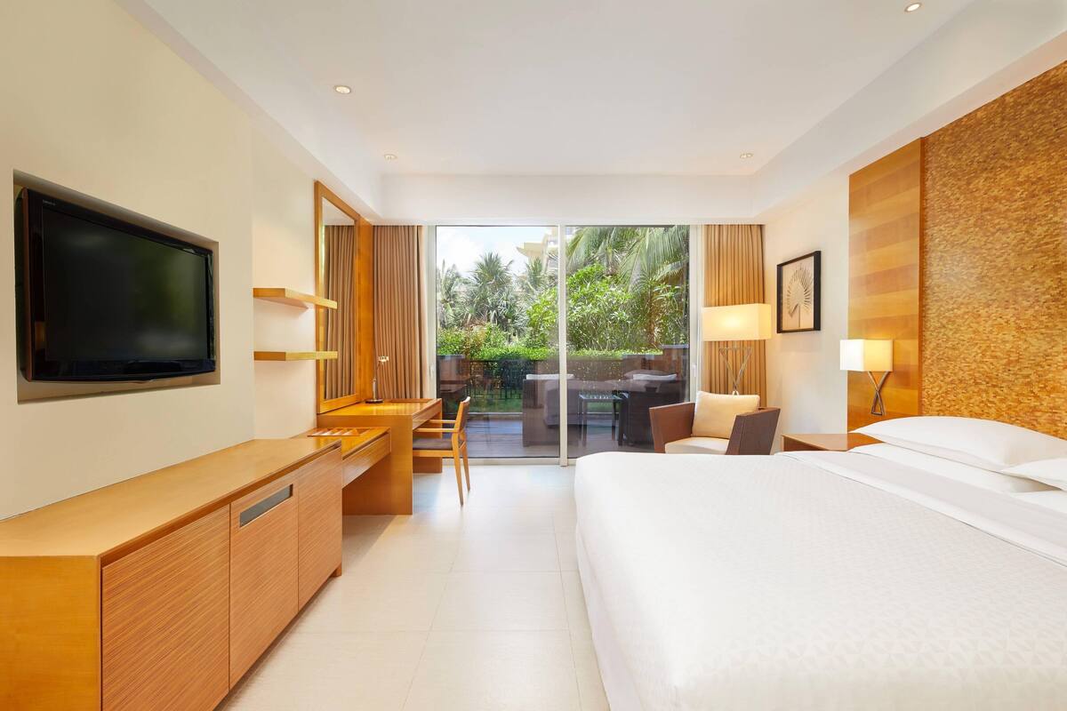 Una habitación de hotel moderna con una cama grande, un escritorio con televisor de pantalla plana, un área de descanso con una mesa y sillas, y vistas al exterior.