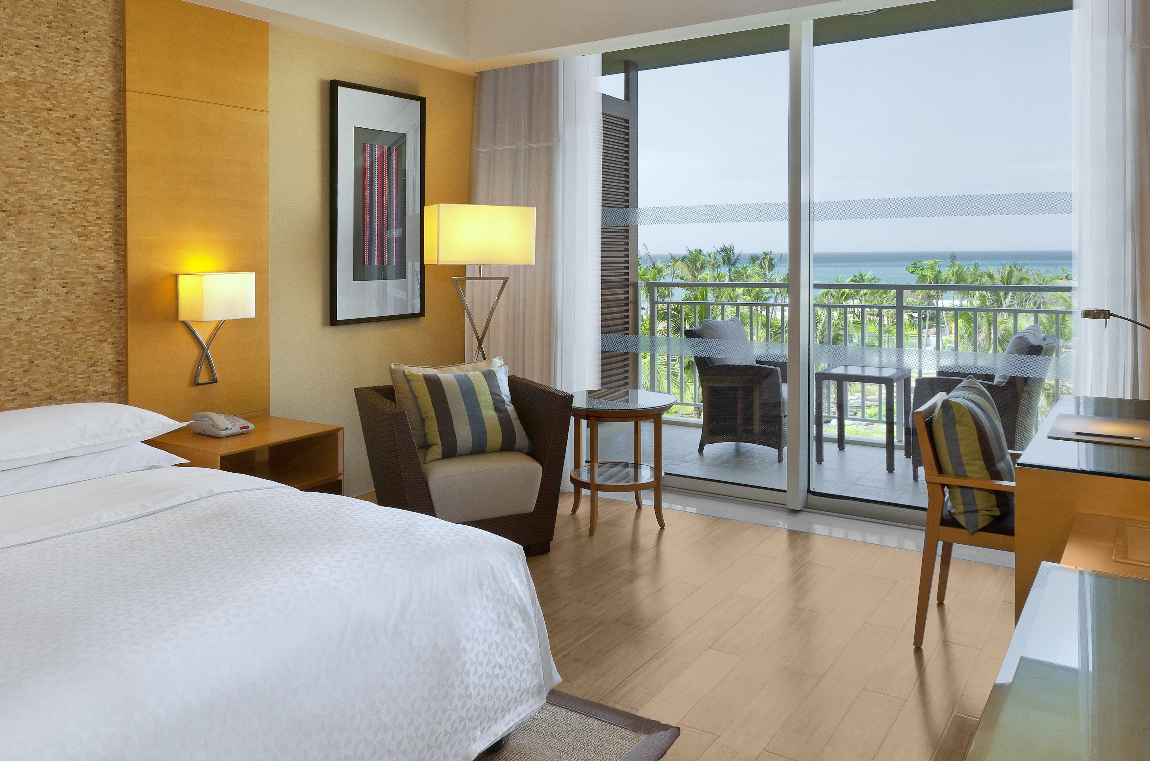 A hotel room with a bed, two bedside tables with lamps, a chair, a small table, and a balcony with a view of the ocean.