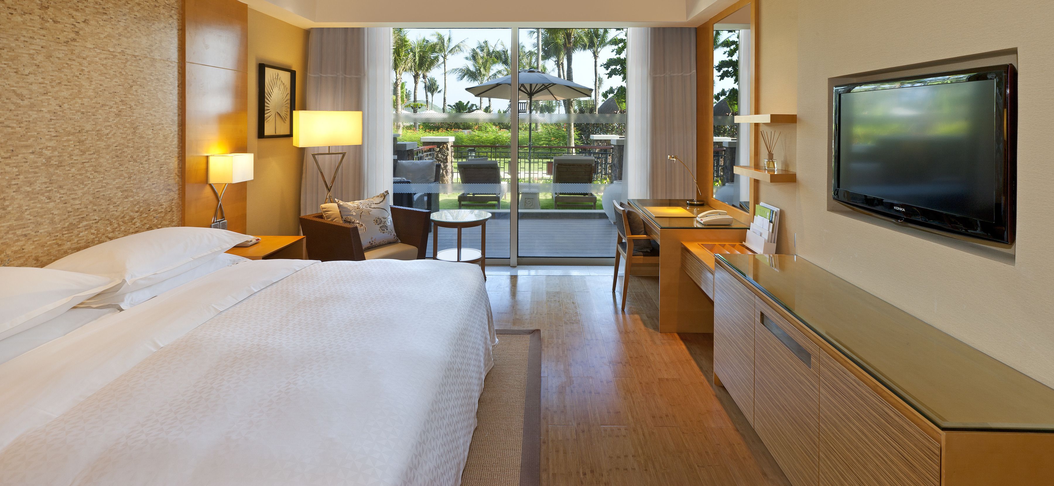 A hotel room with a large bed, a desk with a TV, a chair, and a view of the outdoor patio with palm trees.