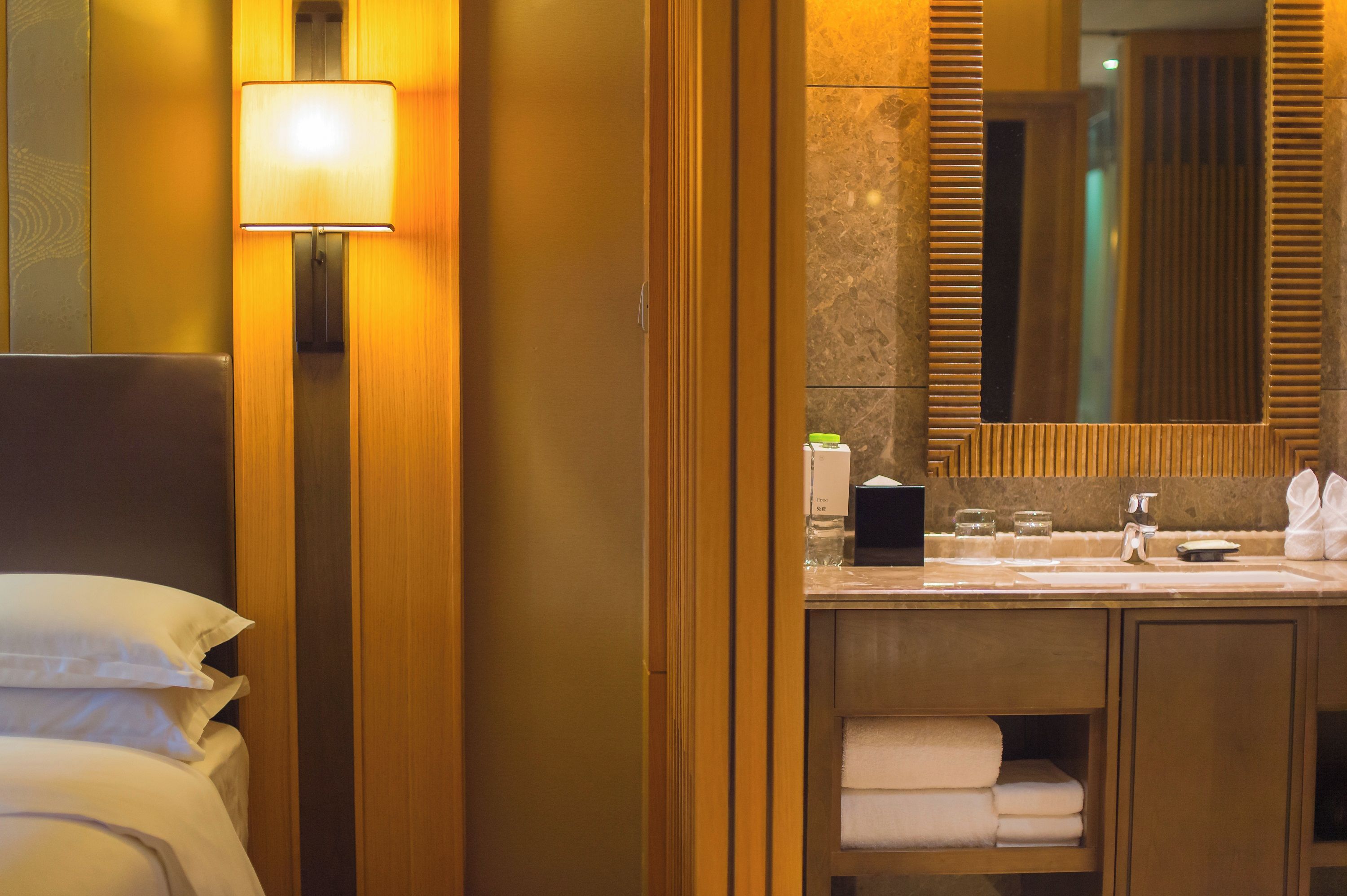 A hotel bathroom with a wooden vanity, a large mirror, and a bed with white linens.