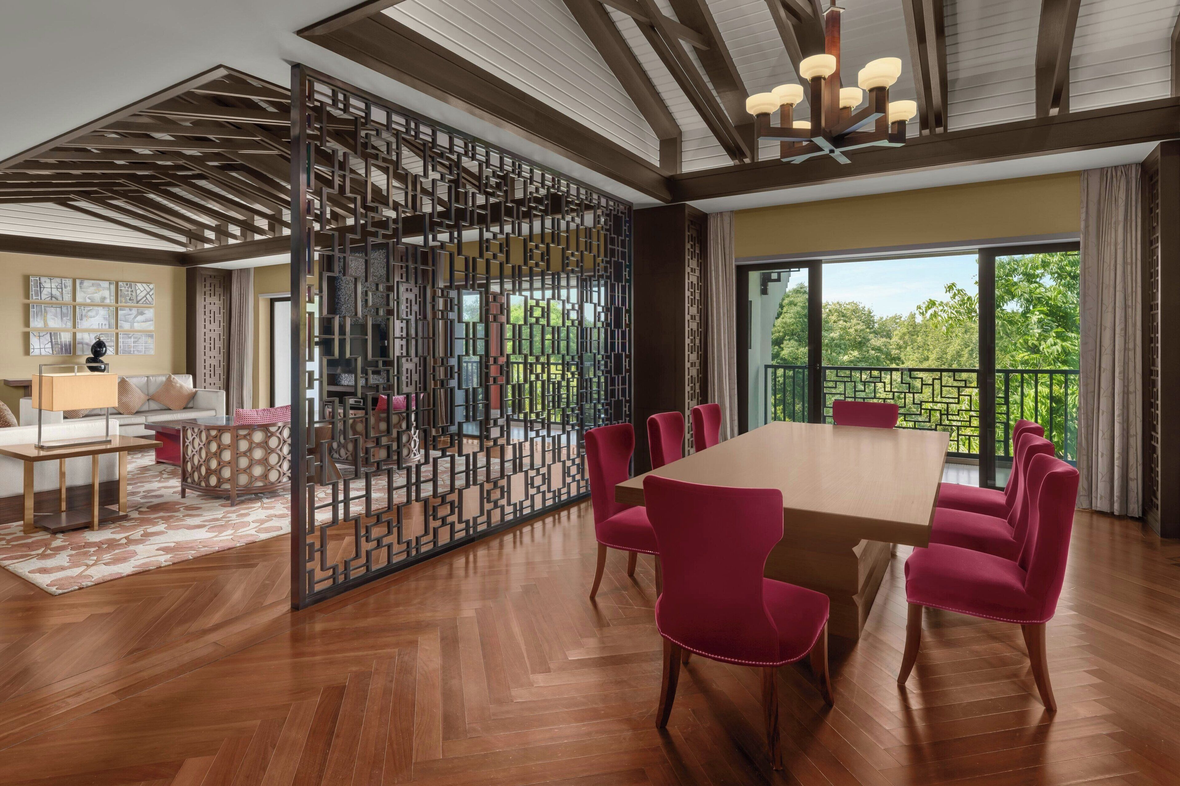 A spacious dining area with a large wooden table and matching chairs, a decorative partition, and a view of greenery through large windows.