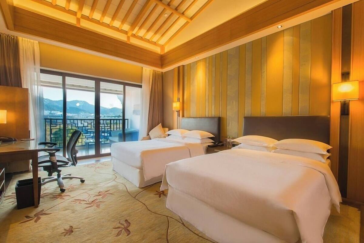 A hotel room with two beds, a desk, a chair, and a large window offering a view of mountains.