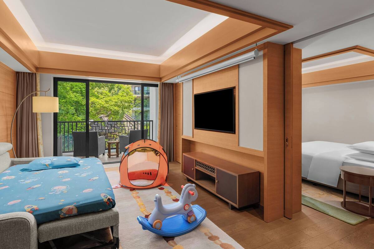 A hotel room with a bed, a TV, a playpen, and a balcony with outdoor furniture.