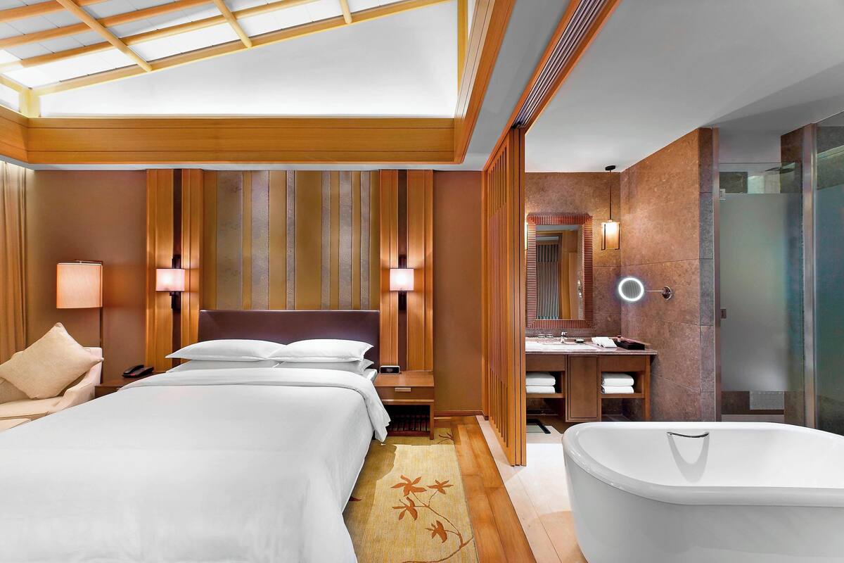 A hotel room with a large bed, a sitting area, a bathroom with a bathtub, and a shower area.