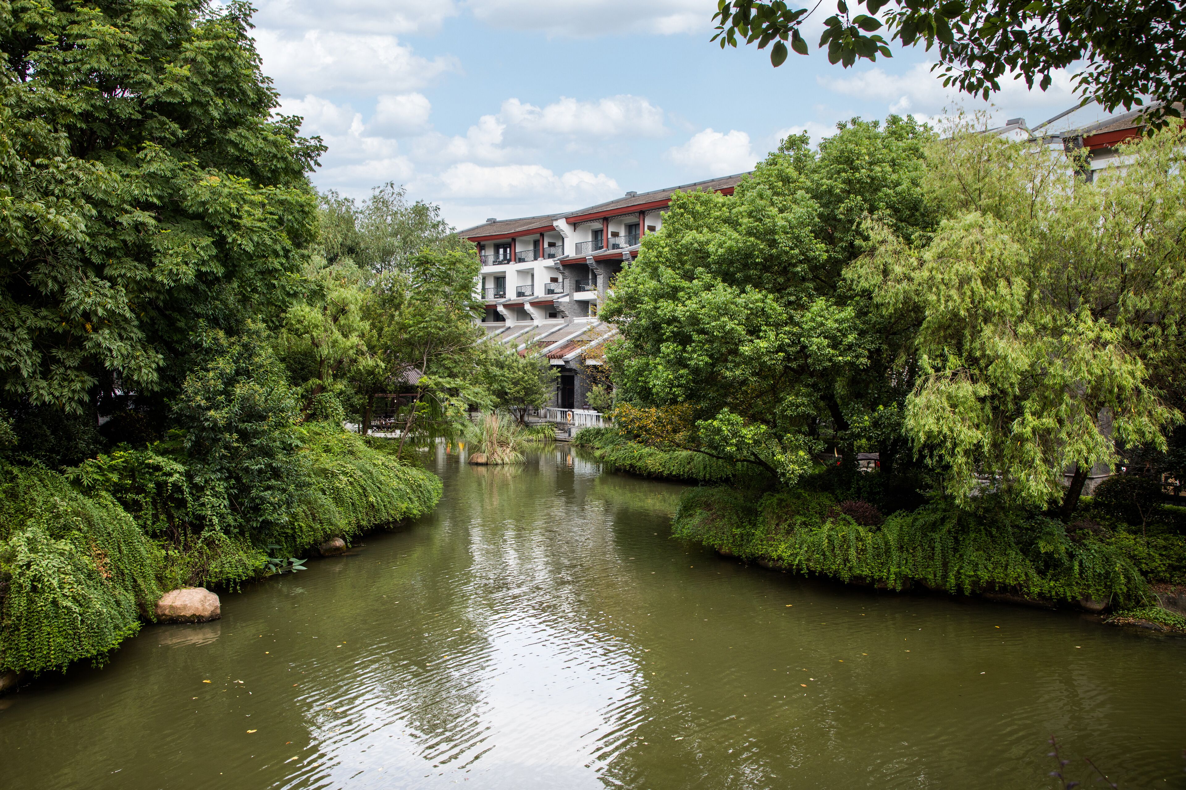 Une rivière bordée d’une végétation luxuriante de chaque côté, un bâtiment avec un toit rouge en arrière-plan et un ciel dégagé.