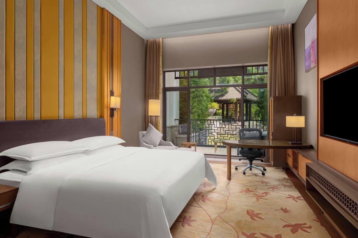 A hotel room with a bed, desk, TV, and a view of a garden through a large window.
