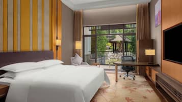 A hotel room with a bed, desk, TV, and a view of a garden through a large window.