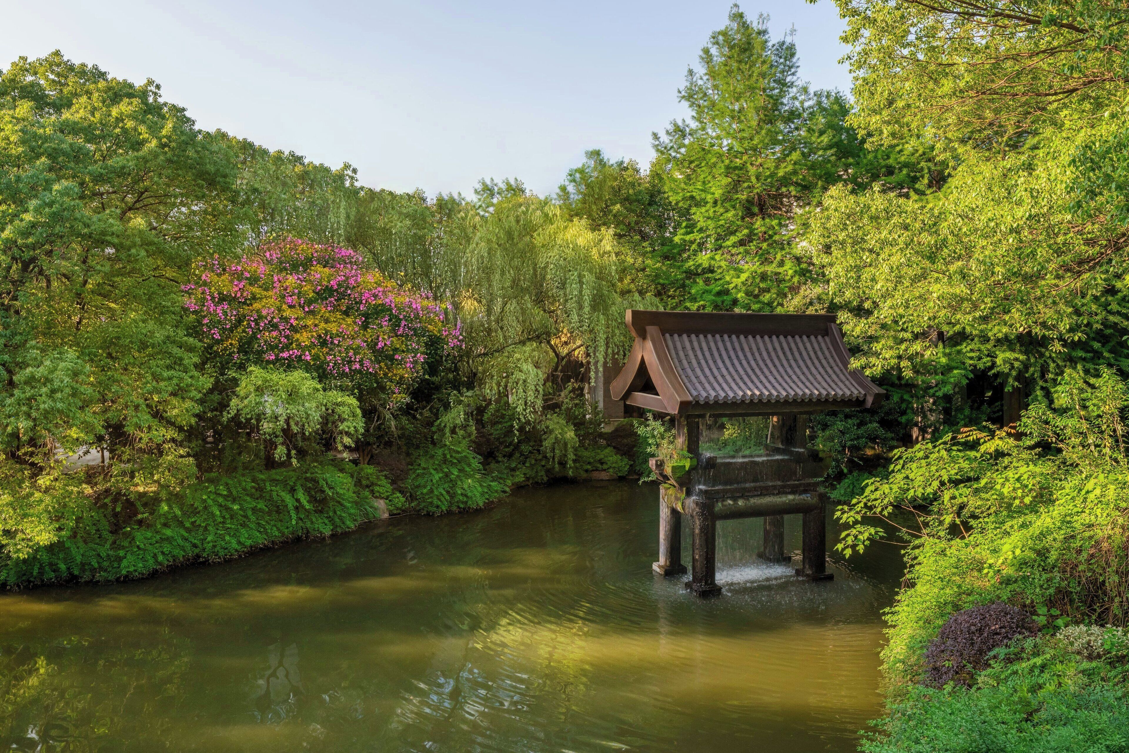 A traditional Japanese-style pavilion over a calm river, surrounded by lush greenery and flowering plants.