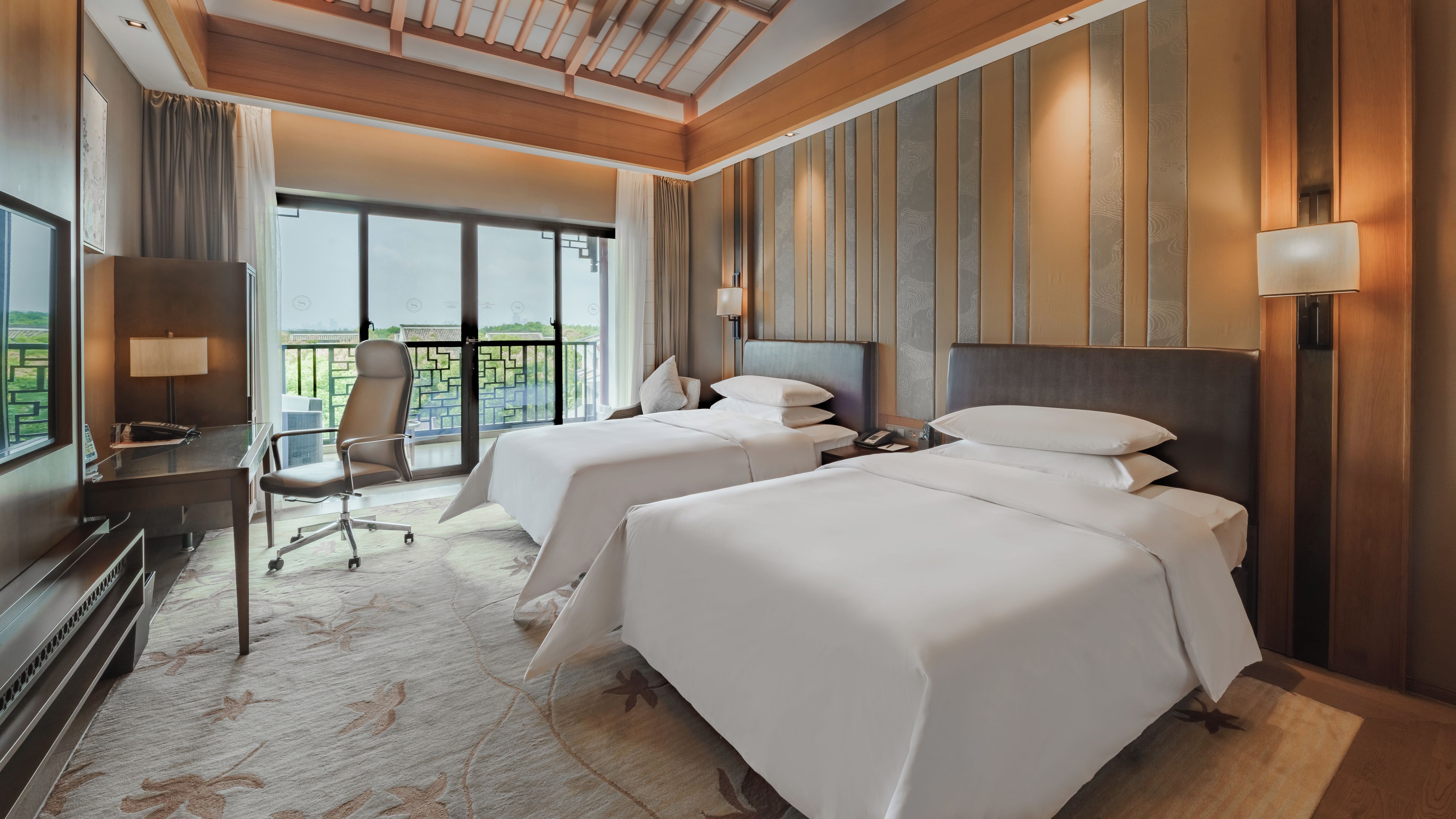 A hotel room with two beds, a desk, a chair, and a balcony with a view.