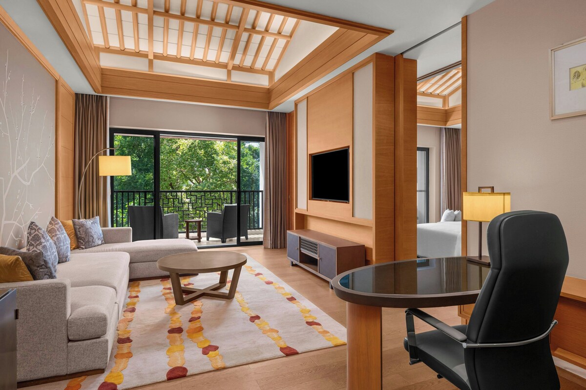 A modern hotel room with a sofa, a round coffee table, a TV, and a balcony with outdoor furniture.