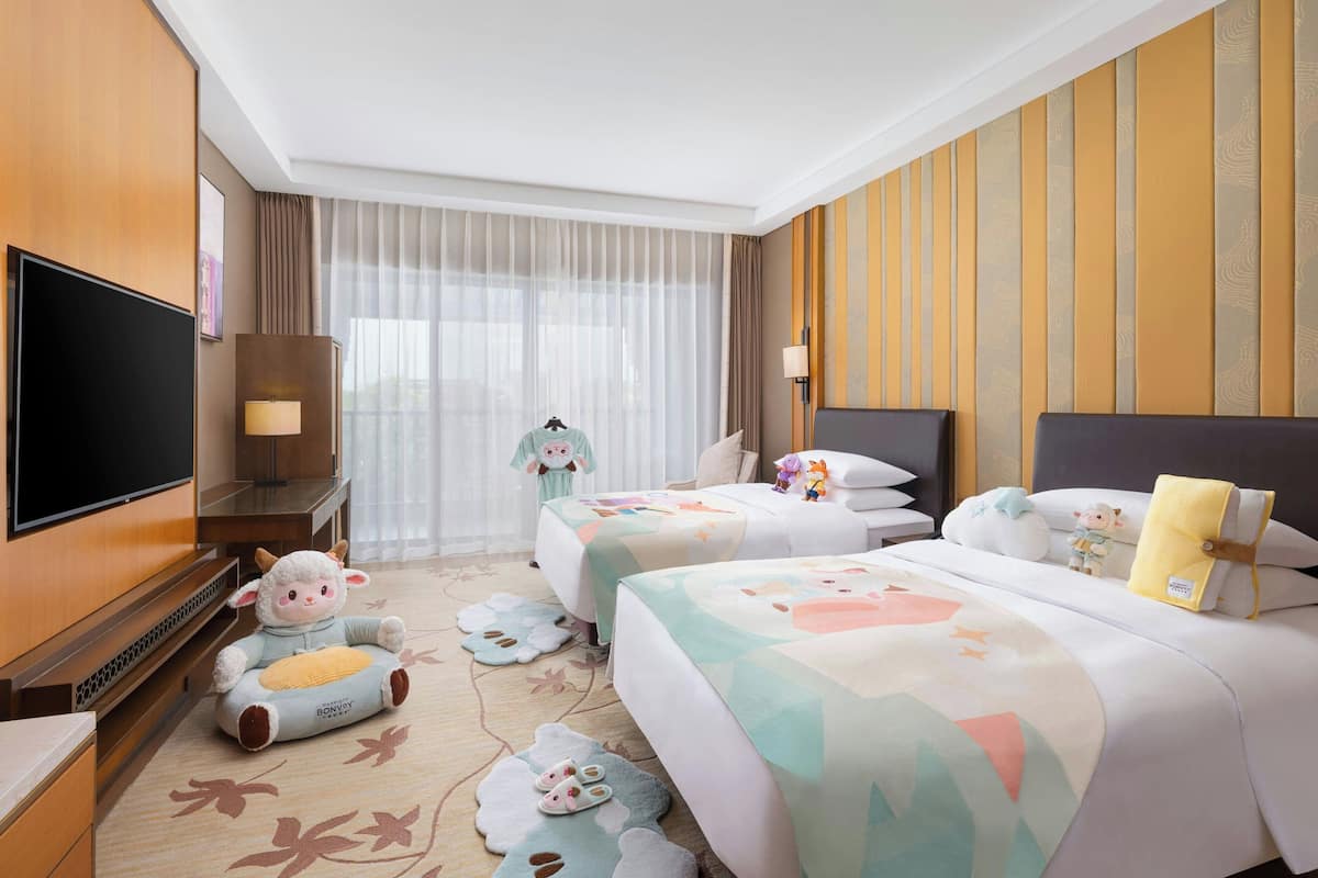 A hotel room with a TV, two beds, a sofa, a TV stand, a lamp, and a window with curtains.
